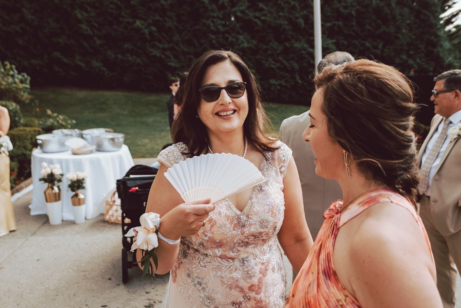 Two women in floral dresses chat at an outdoor wedding, one wearing sunglasses and holding a white hand fan.