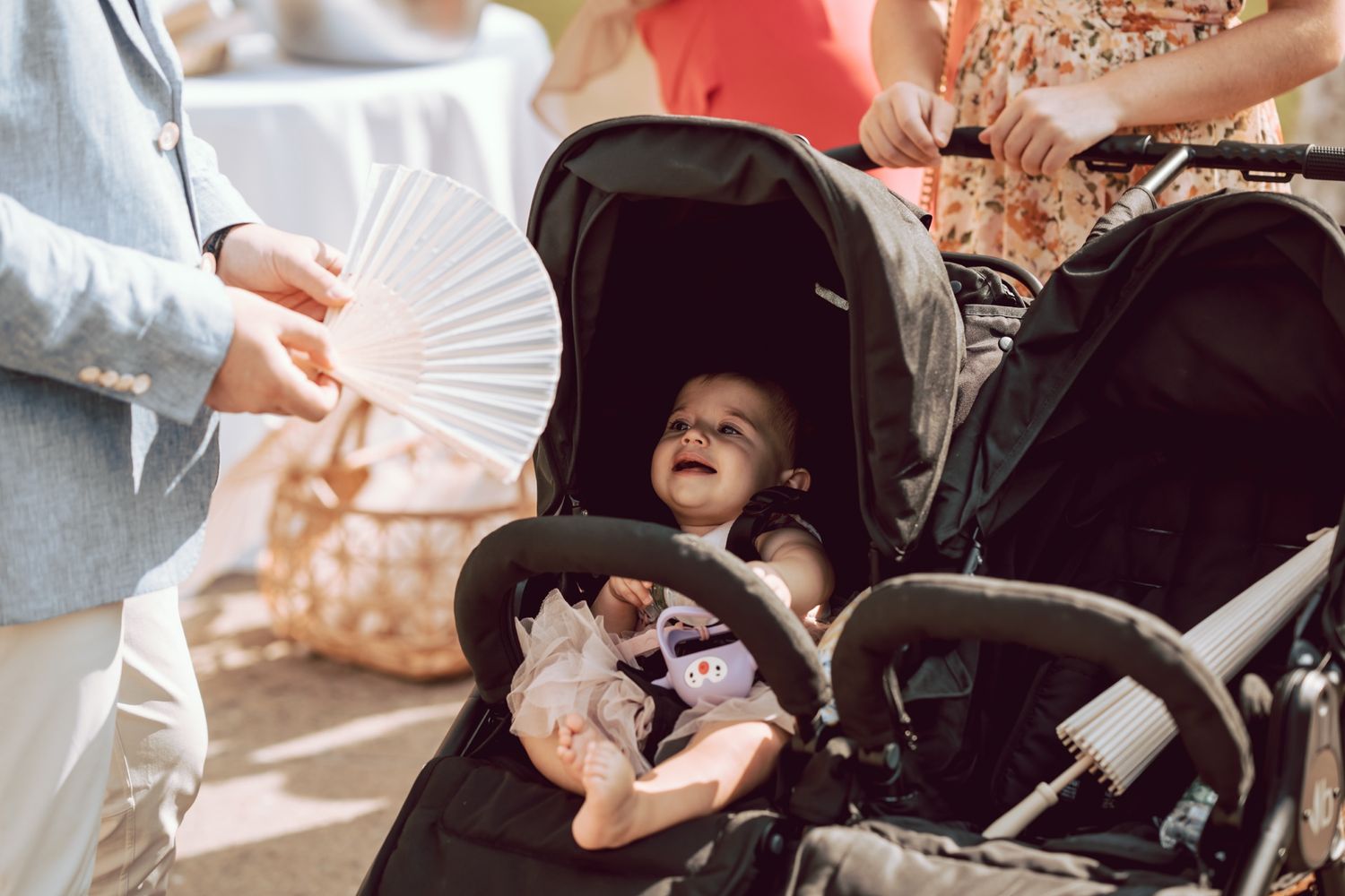 Happy baby in black stroller smiles at white hand fan at outdoor wedding celebration.
