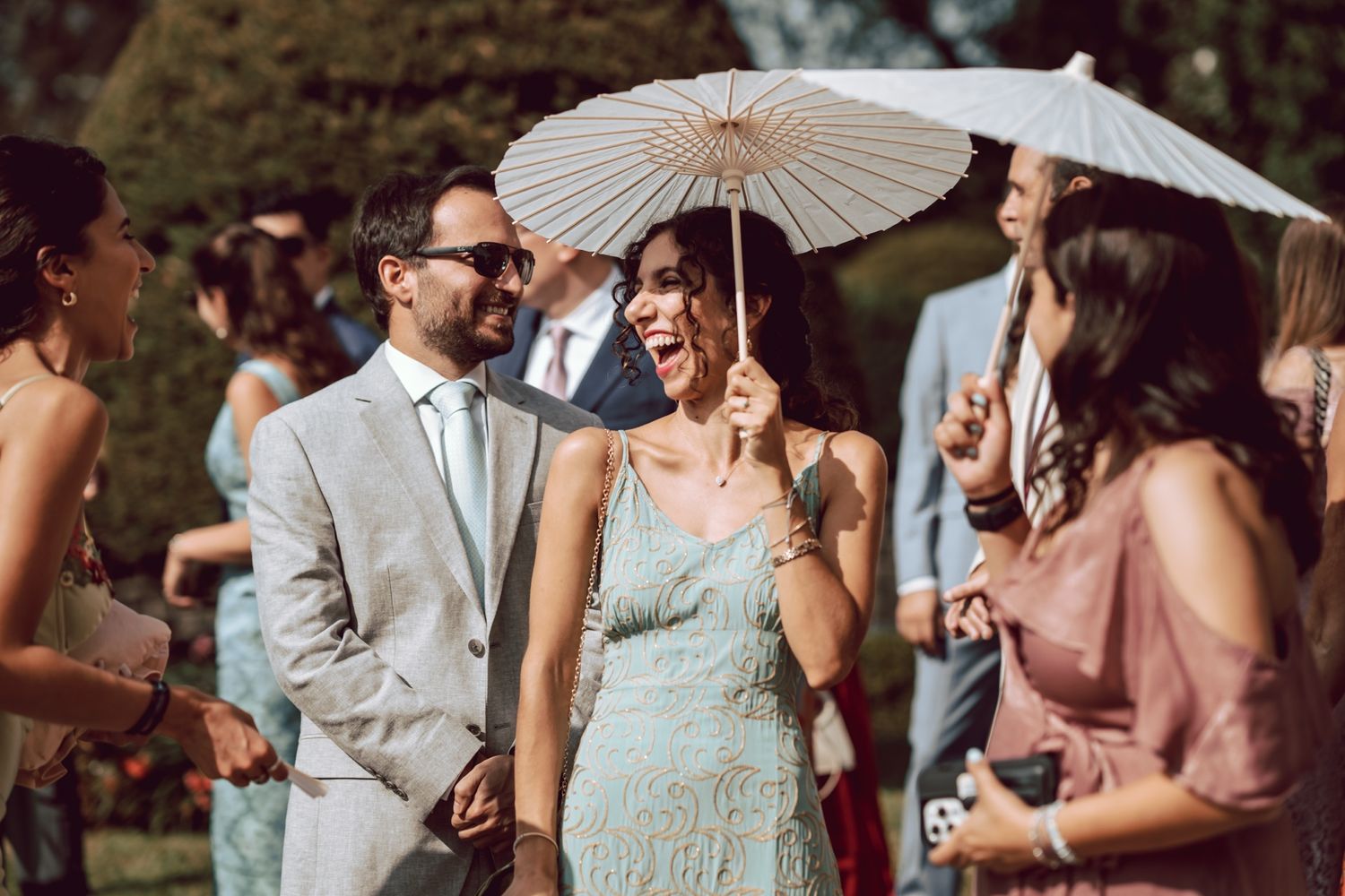 Elegant wedding guests laughing outdoors, woman in mint dress holding white parasol beside man in gray suit.