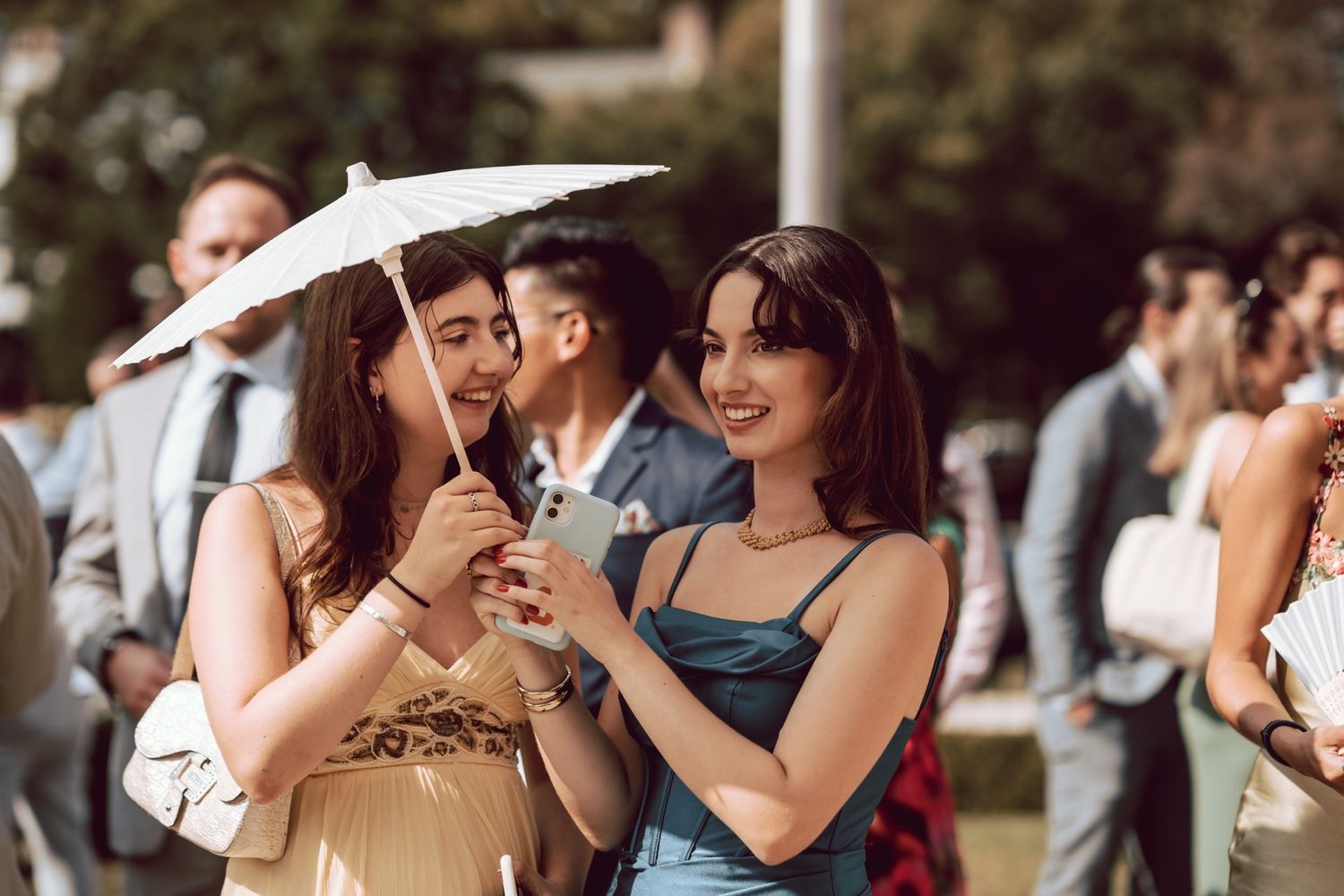 Two women smile at a phone at an outdoor wedding, one holding a white parasol.
