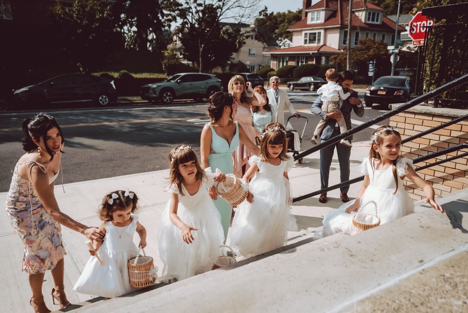 Flower girls in white dresses walk down steps at a sunny outdoor wedding ceremony.
