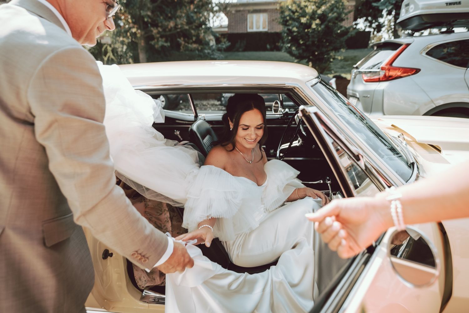 Bride in white gown being helped out of a vintage car by wedding guests on her special day.