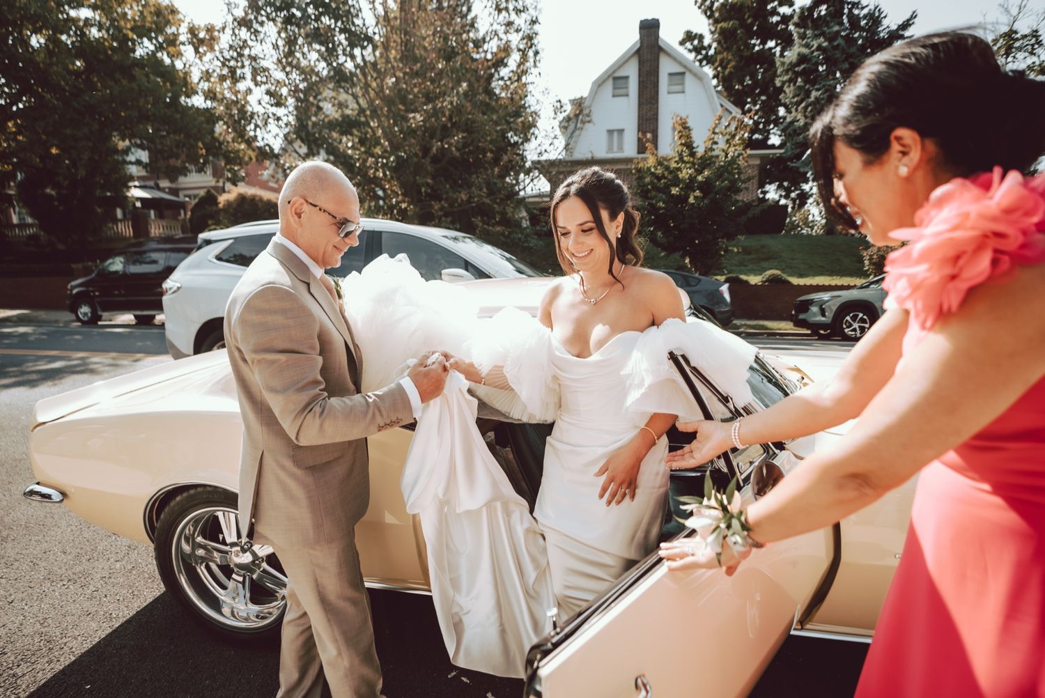 Bride in white gown exits vintage cream car assisted by man in tan suit and woman in pink dress outdoors.