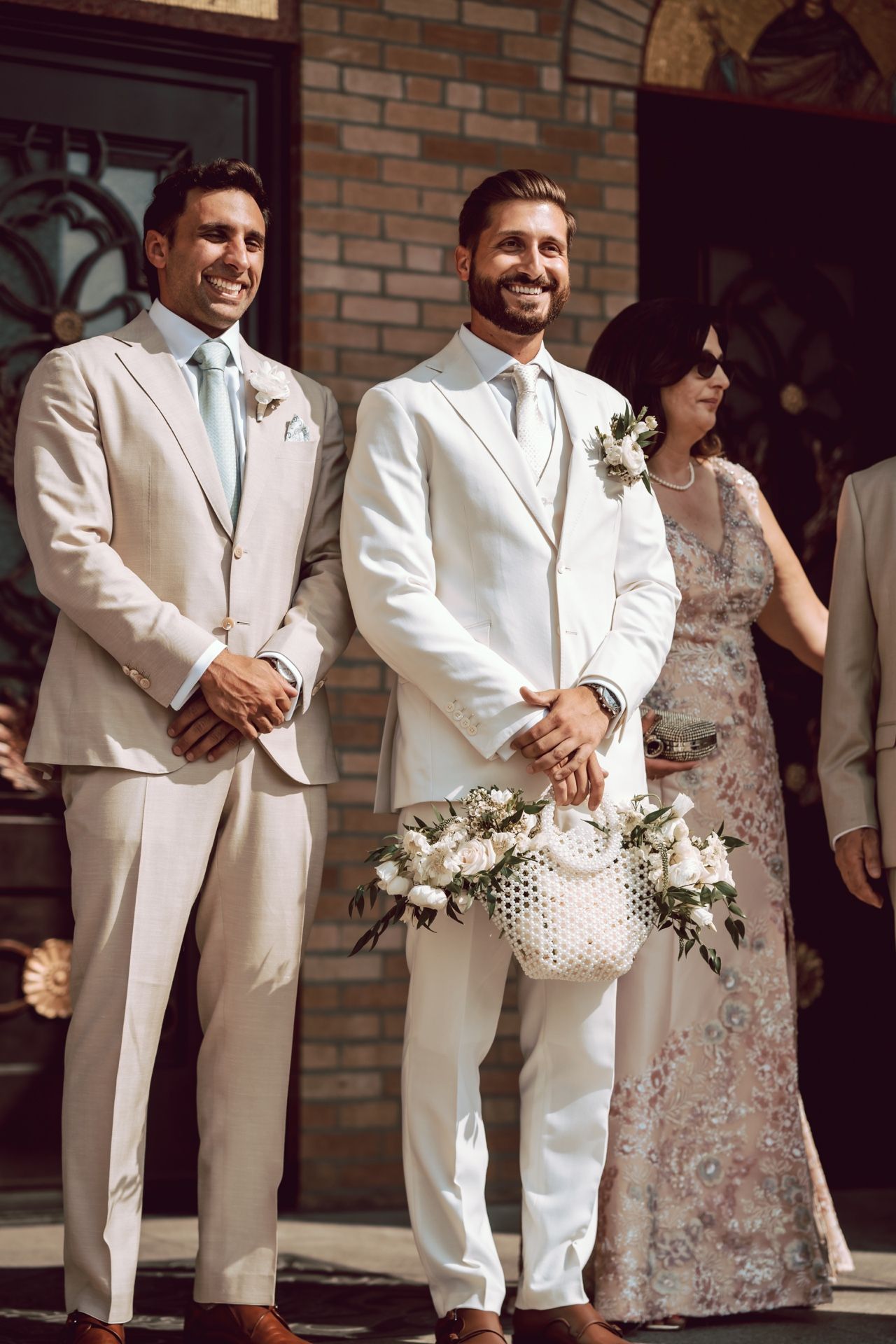 Groom in white suit smiling with groomsman in beige suit holding floral basket at wedding ceremony.