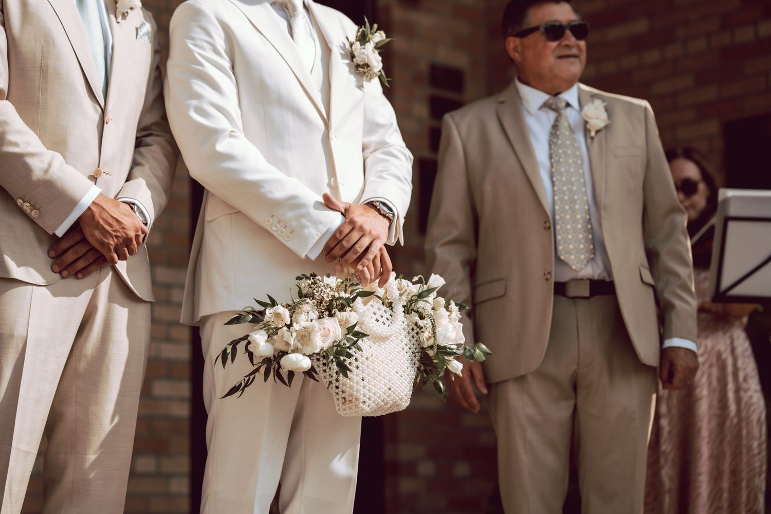Men in beige and white suits at a wedding ceremony, one holding a white floral basket with greenery.