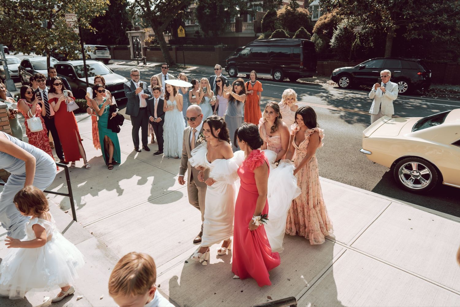 Wedding party celebrating outdoors with guests cheering, bride in white gown, bridesmaids in colorful dresses on sunny day.