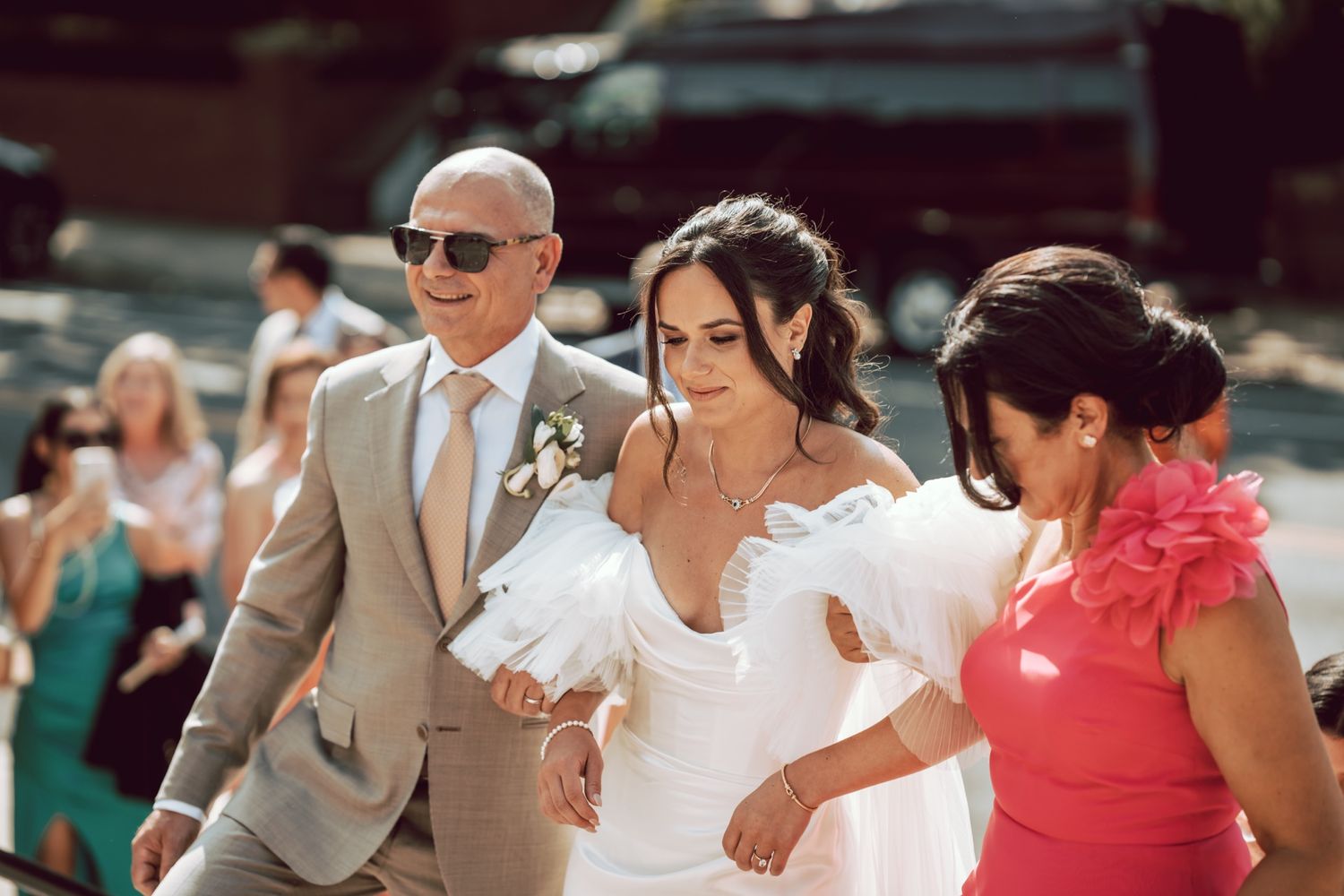 Bride in white gown walks down aisle escorted by man in tan suit and woman in pink dress at outdoor wedding.