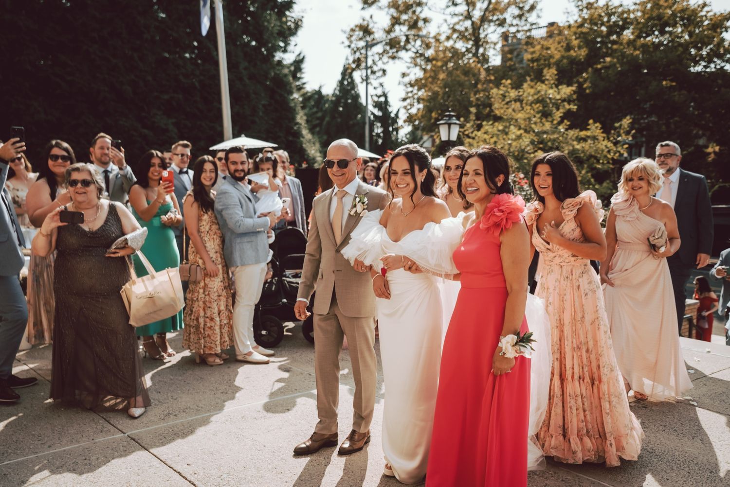 Bride in white dress walks with wedding party and guests gathered outdoors on sunny day.