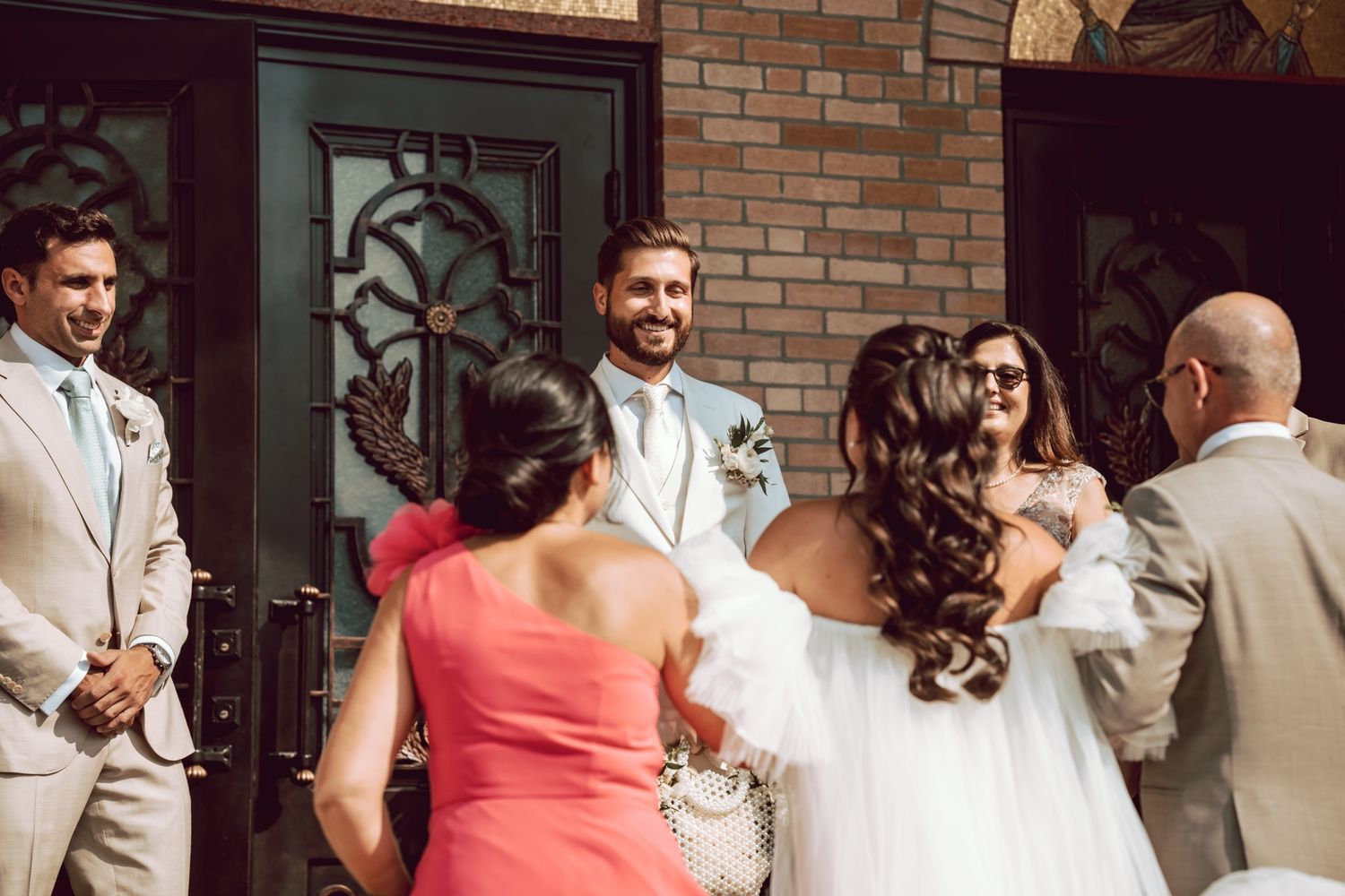 Bride and groom greeting wedding guests outside a church door with bridesmaids in coral dresses.