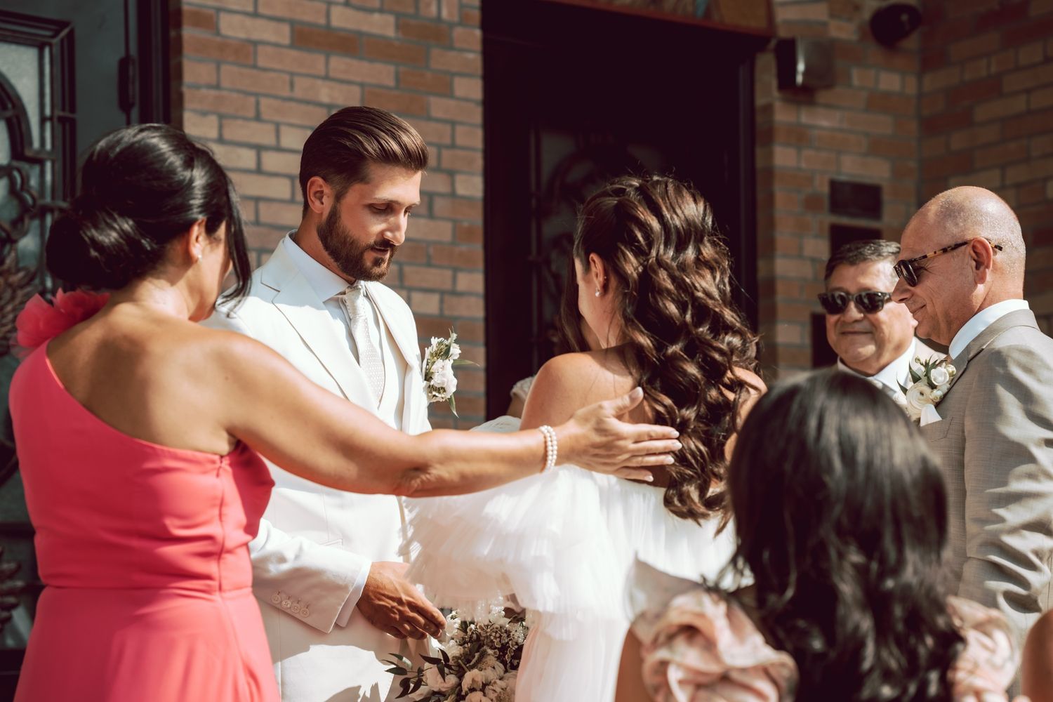 Bride and groom greeting wedding guests outside a brick building, exchanging warm embraces in elegant attire.
