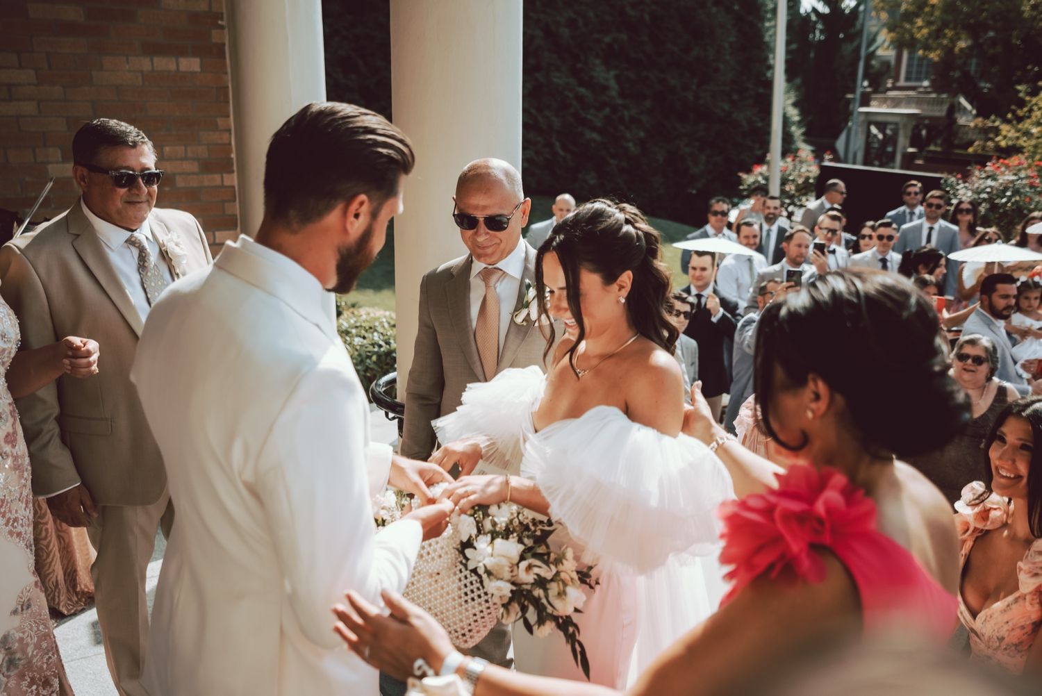 Bride in white ruffled gown holds bouquet while greeting groom outdoors at elegant wedding ceremony.