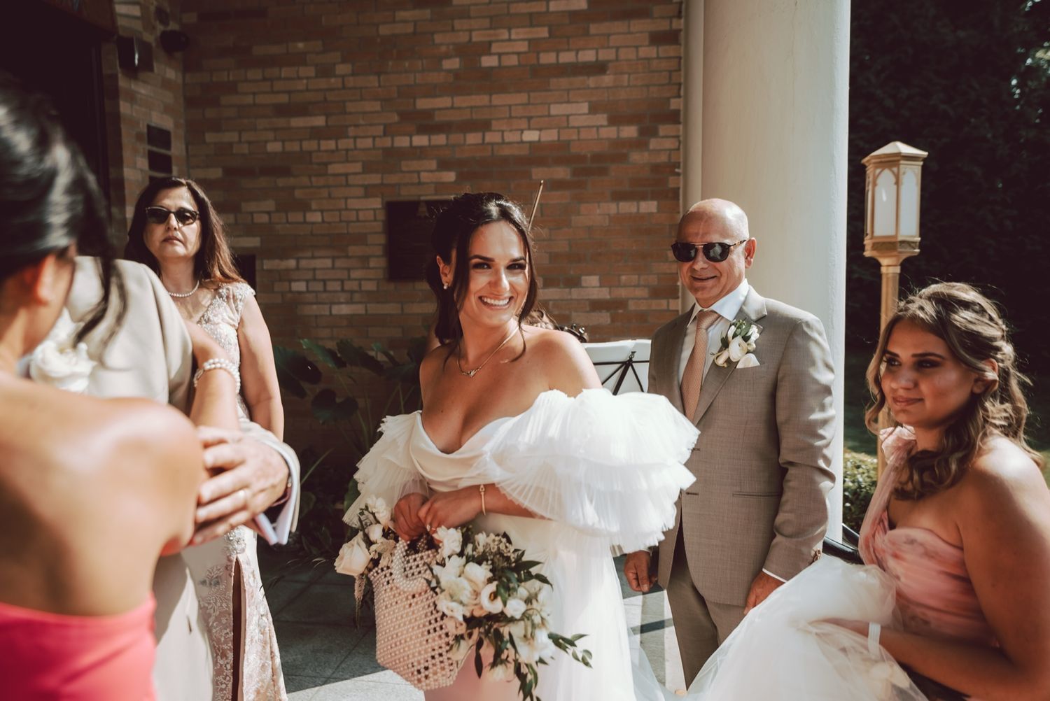 Smiling bride in white off-shoulder gown holding bouquet surrounded by wedding guests at outdoor ceremony.