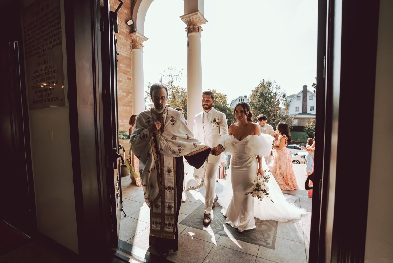 Bride and groom exit church with officiant in ornate robes during outdoor wedding ceremony.