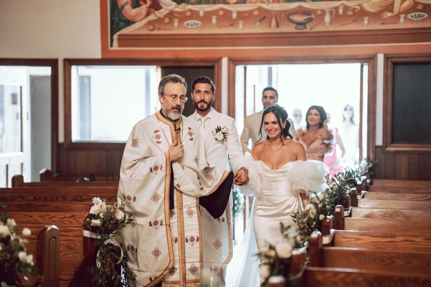 Bride and groom walking down the aisle with an Orthodox priest during a church wedding ceremony.