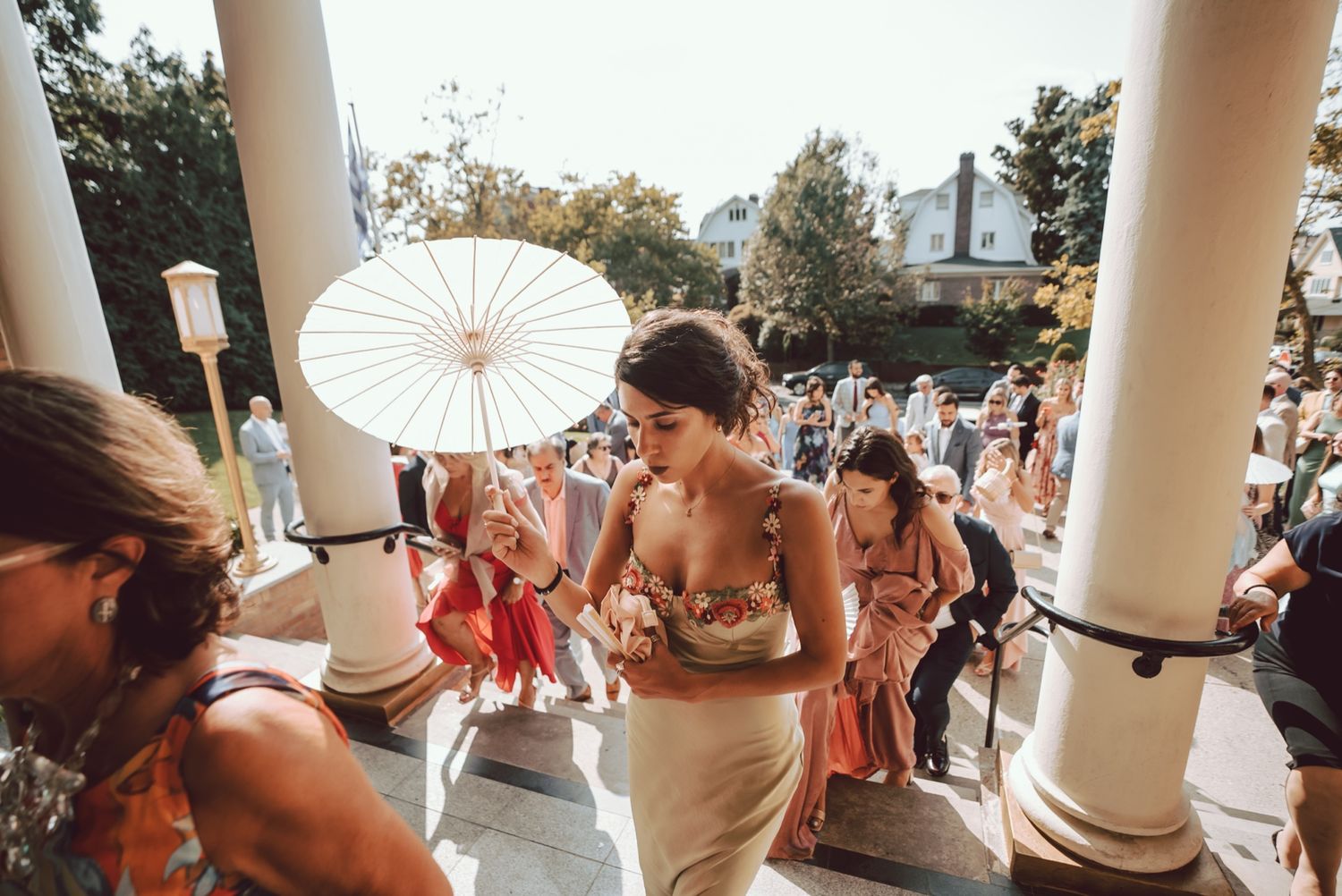 Bride holding white parasol descends steps at outdoor wedding ceremony with guests and columns.