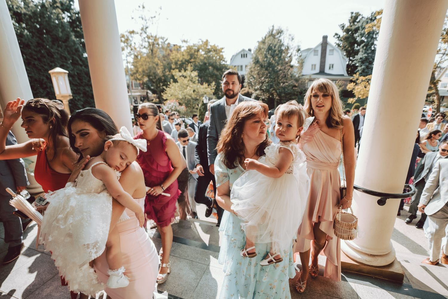 Wedding guests descend stairs, including women in pink dresses carrying children in white outfits, at an outdoor ceremony.