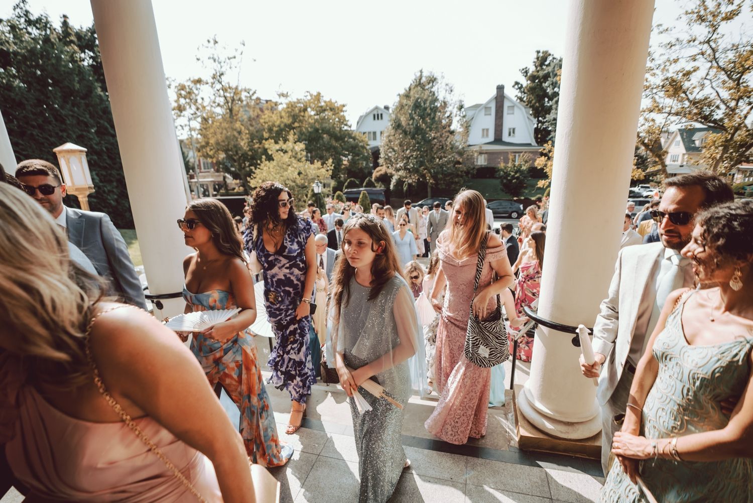 Wedding guests in colorful attire socializing on a porch with white columns at an outdoor event.