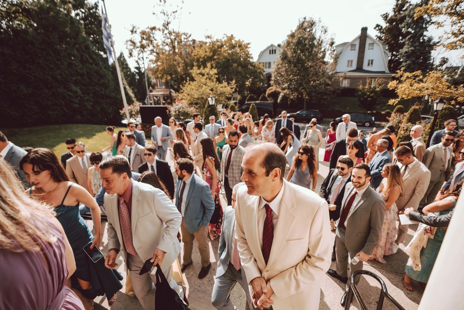 Elegantly dressed wedding guests mingling outdoors on a sunny day near a classic white house.