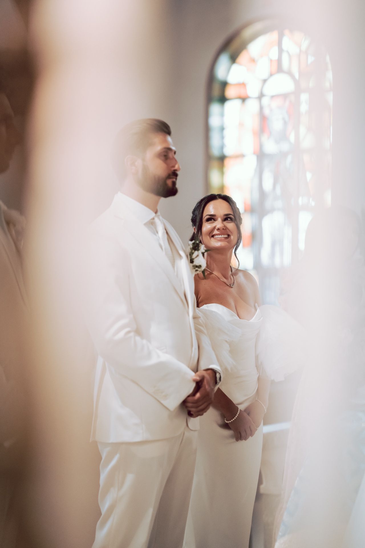 Bride and groom in matching white attire hold hands during church wedding ceremony by stained glass.