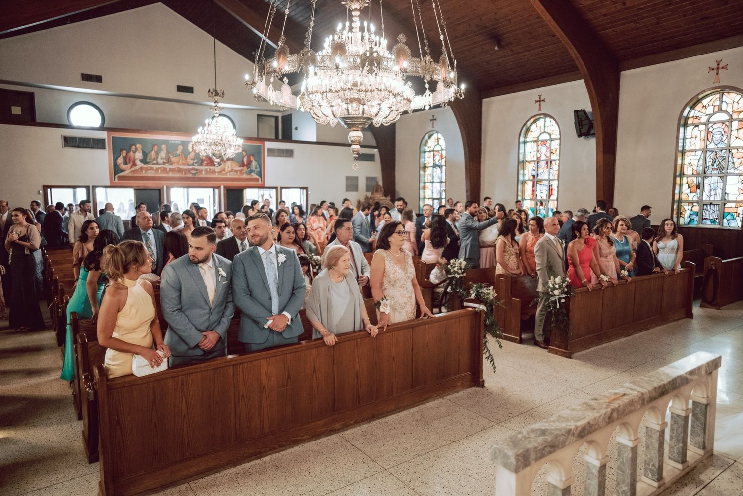 Guests standing in wooden pews during a wedding ceremony inside an elegant church with stained glass windows and chandelier.