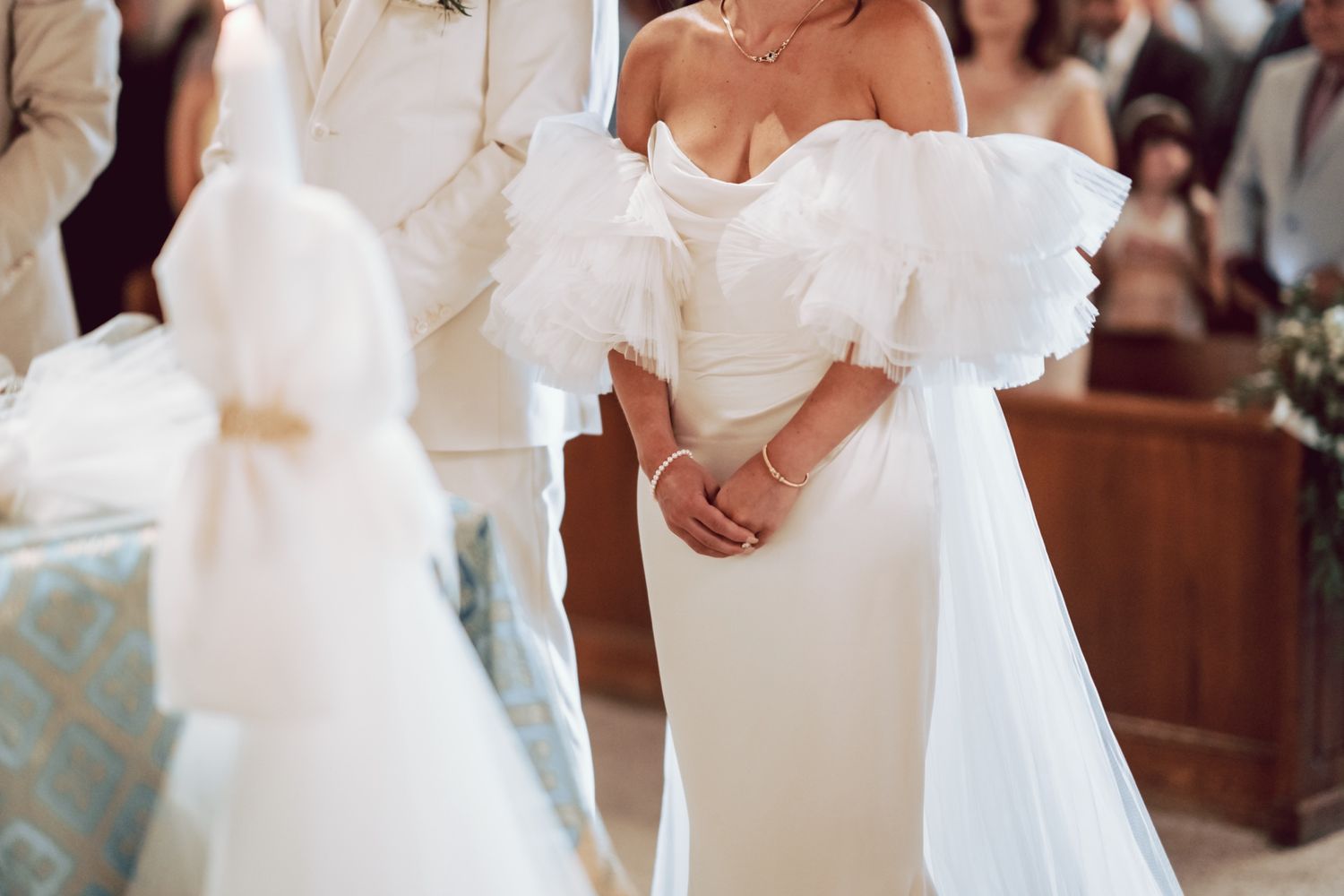 Bride in elegant off-shoulder white ruffle wedding gown standing at church altar during ceremony.