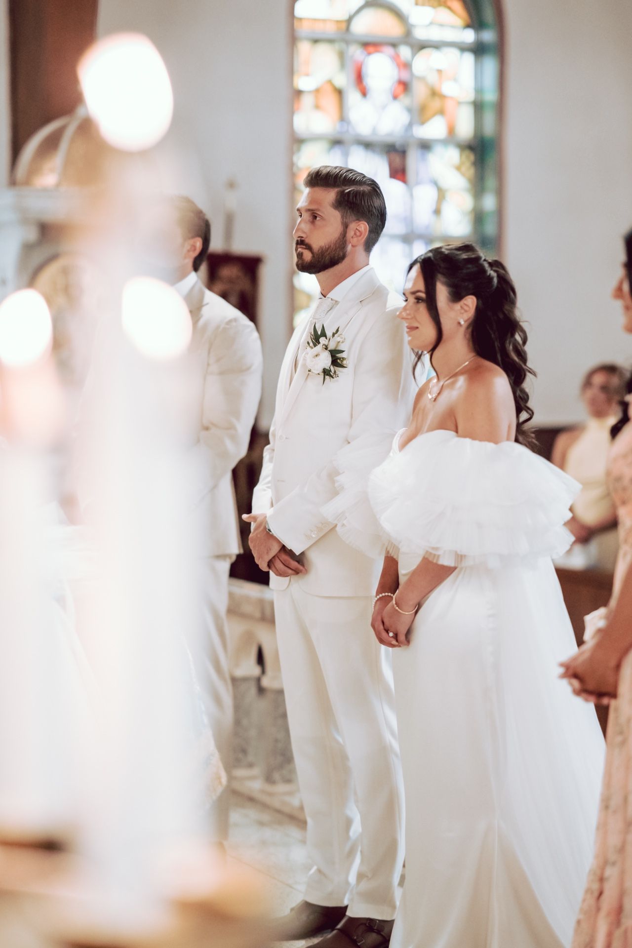 Bride and groom standing at the altar during a church wedding ceremony, dressed in white attire with candles in the foreground.