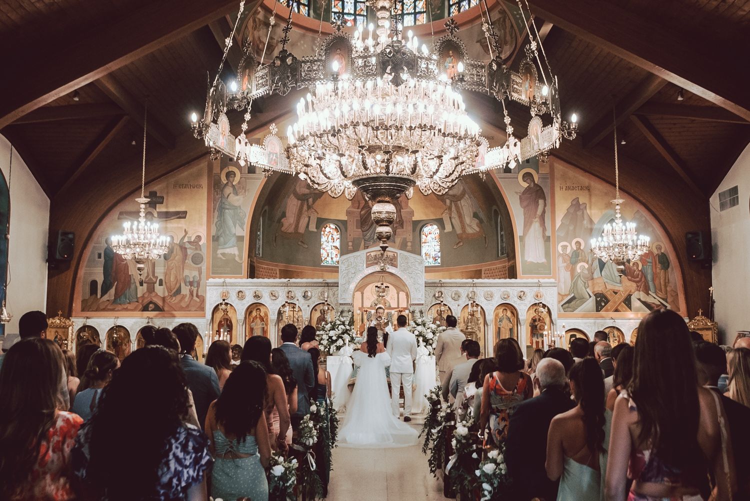 Grand Orthodox church wedding ceremony with guests seated in pews beneath ornate chandeliers.