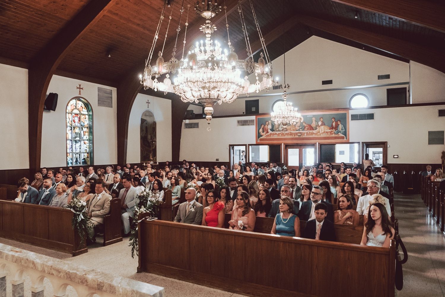 Guests seated in wooden pews of elegant church during wedding ceremony with chandelier.