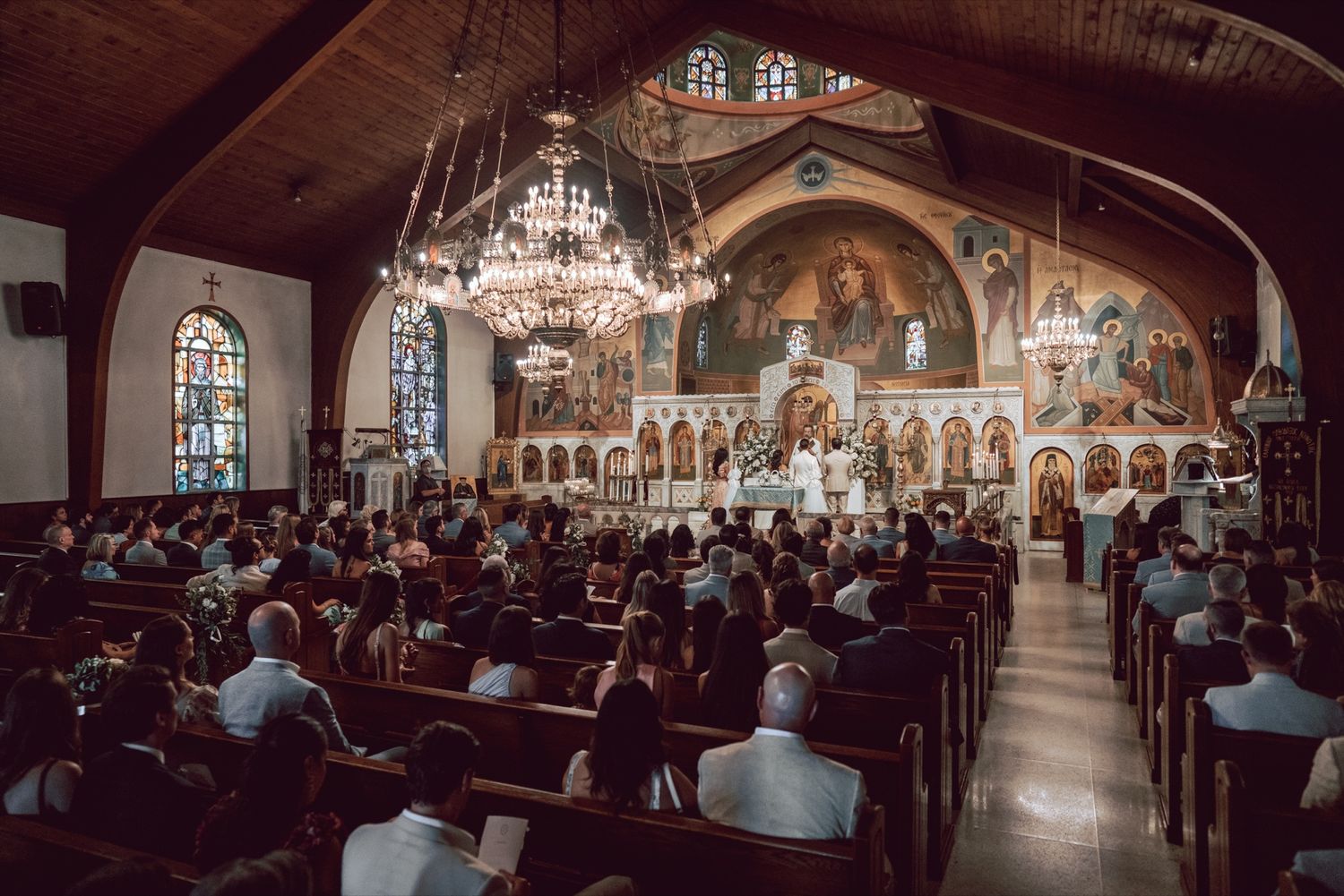 Packed Orthodox church wedding with guests filling wooden pews beneath stunning Byzantine artwork.