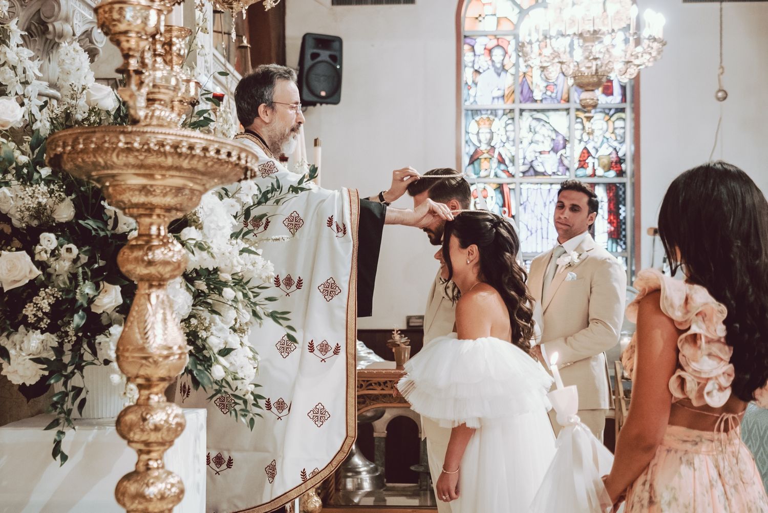 Orthodox wedding ceremony with priest crowning bride and groom at ornate church altar with white floral arrangements.