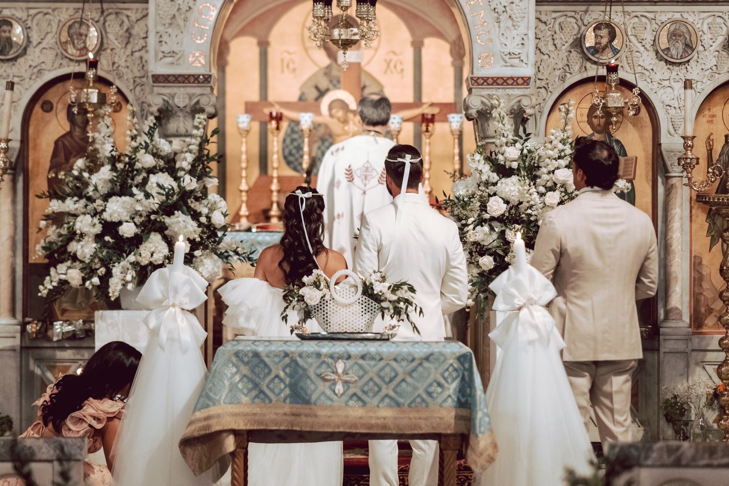 Bride and groom stand before Orthodox altar adorned with white floral arrangements during ceremony.