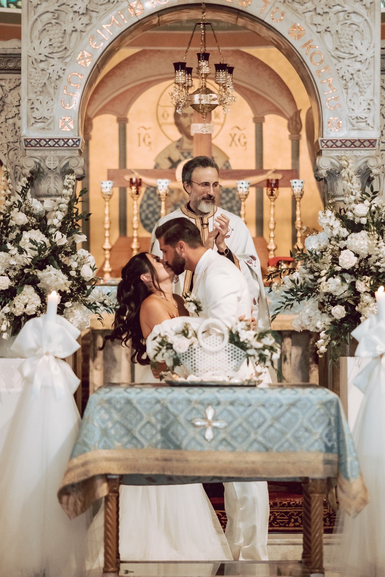 Couple sharing first kiss at Orthodox church wedding altar, officiated by priest amid white floral arrangements.