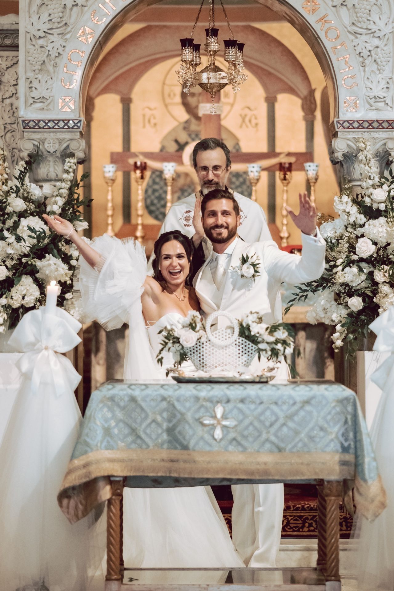 Joyful bride and groom celebrate at the altar of an ornate Orthodox church with white floral arrangements.