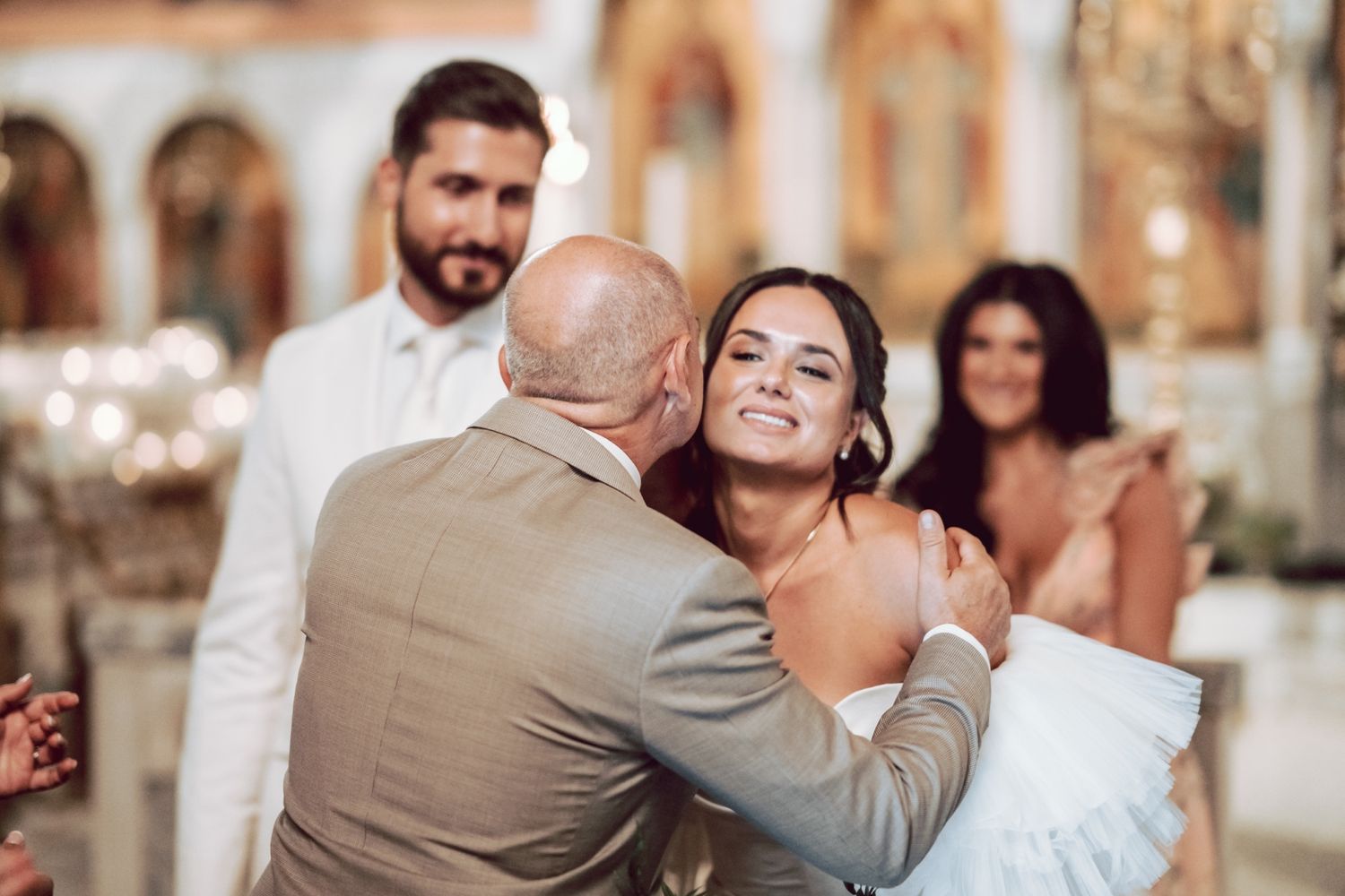Bride smiling joyfully as an older man in a beige suit embraces her during a church wedding ceremony.