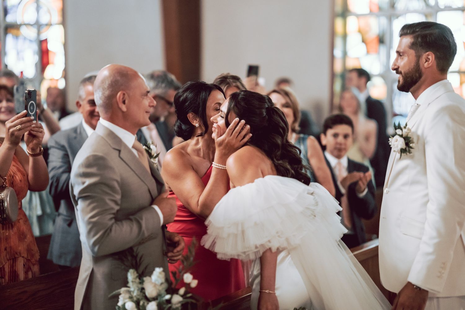 Emotional wedding ceremony moment as bride receives hugs from family members while groom watches in white suit.