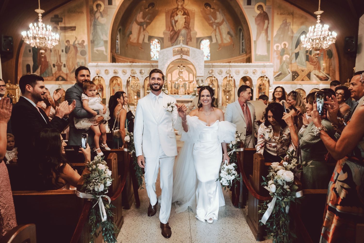 Bride and groom walk down the aisle of an ornate Orthodox church surrounded by cheering guests at their wedding ceremony.