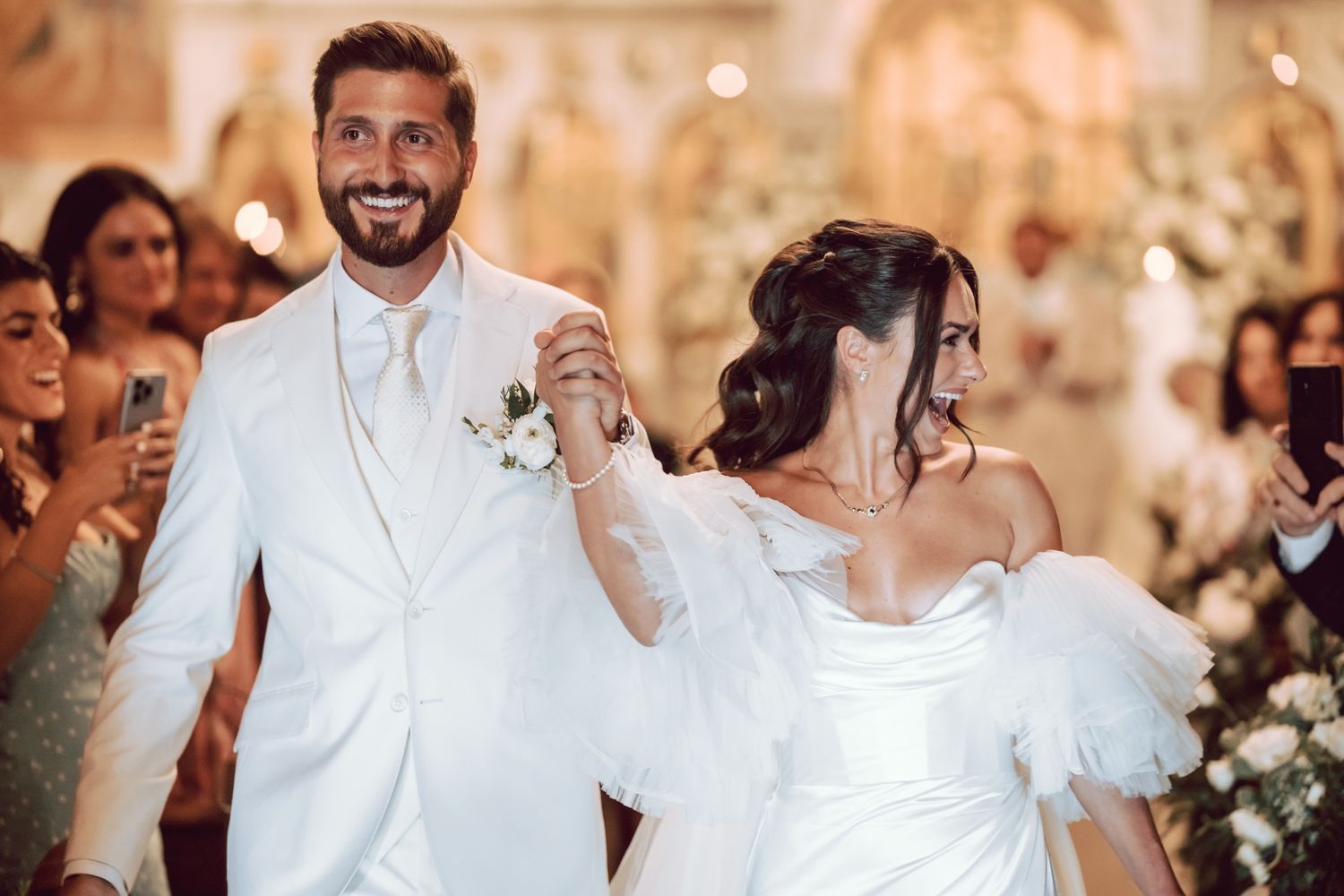 A joyful couple walks down the aisle at their wedding, the groom in white suit and bride in elegant gown.