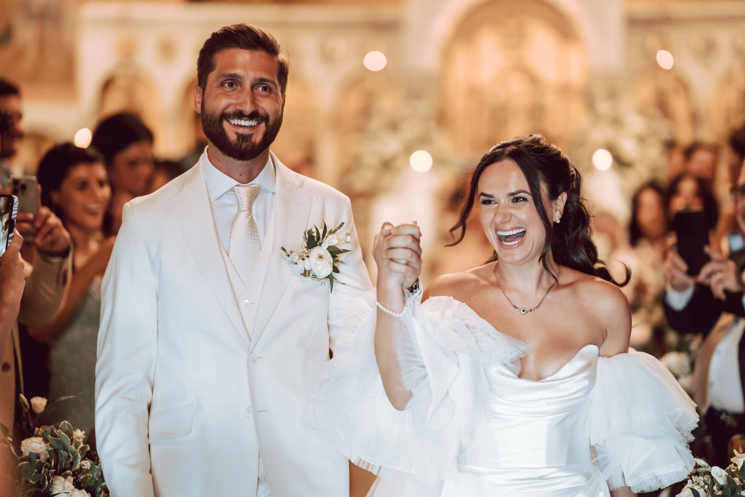 Happy bride and groom in white attire celebrate their wedding ceremony, raising hands joyfully in an elegant church.