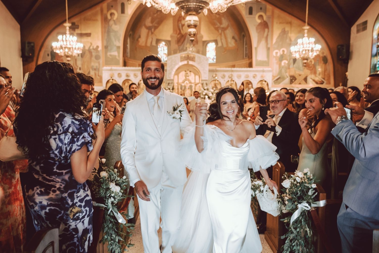 Joyful newlyweds walk down church aisle in white wedding attire, surrounded by cheering guests in elegant ceremony.