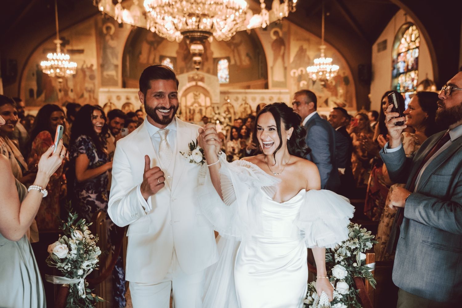 Joyful newlyweds walk down the aisle in an ornate church, groom in white suit, bride in strapless gown, guests cheering.