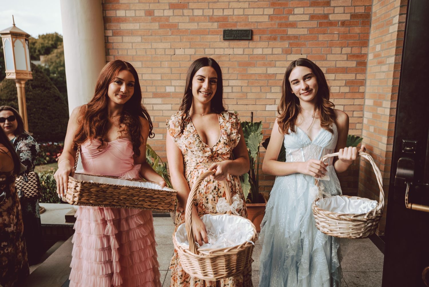 Three bridesmaids in colorful dresses hold decorative baskets outside a brick church at a wedding.