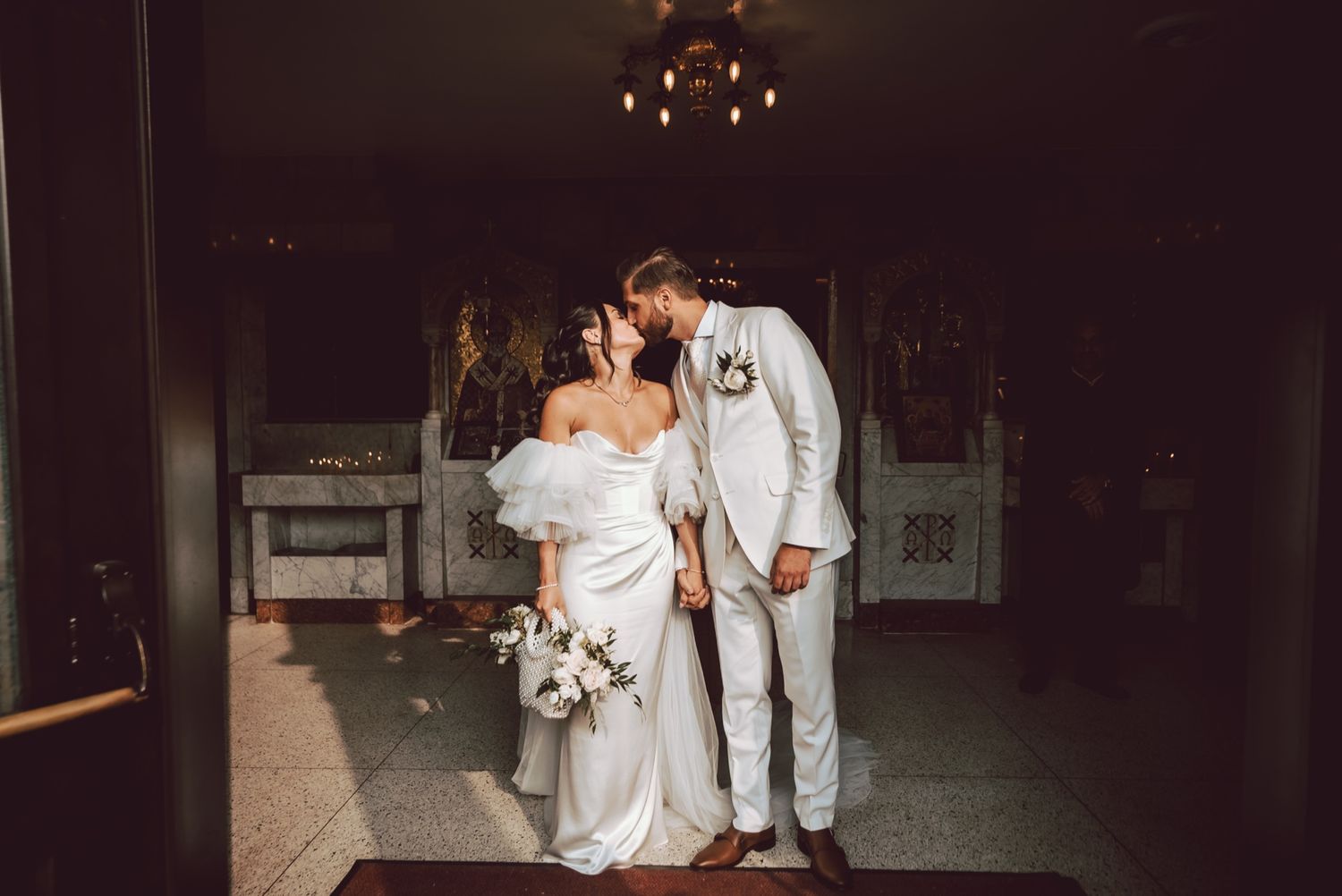 Bride and groom share a romantic kiss in the dimly lit aisle of a beautiful church after their ceremony.