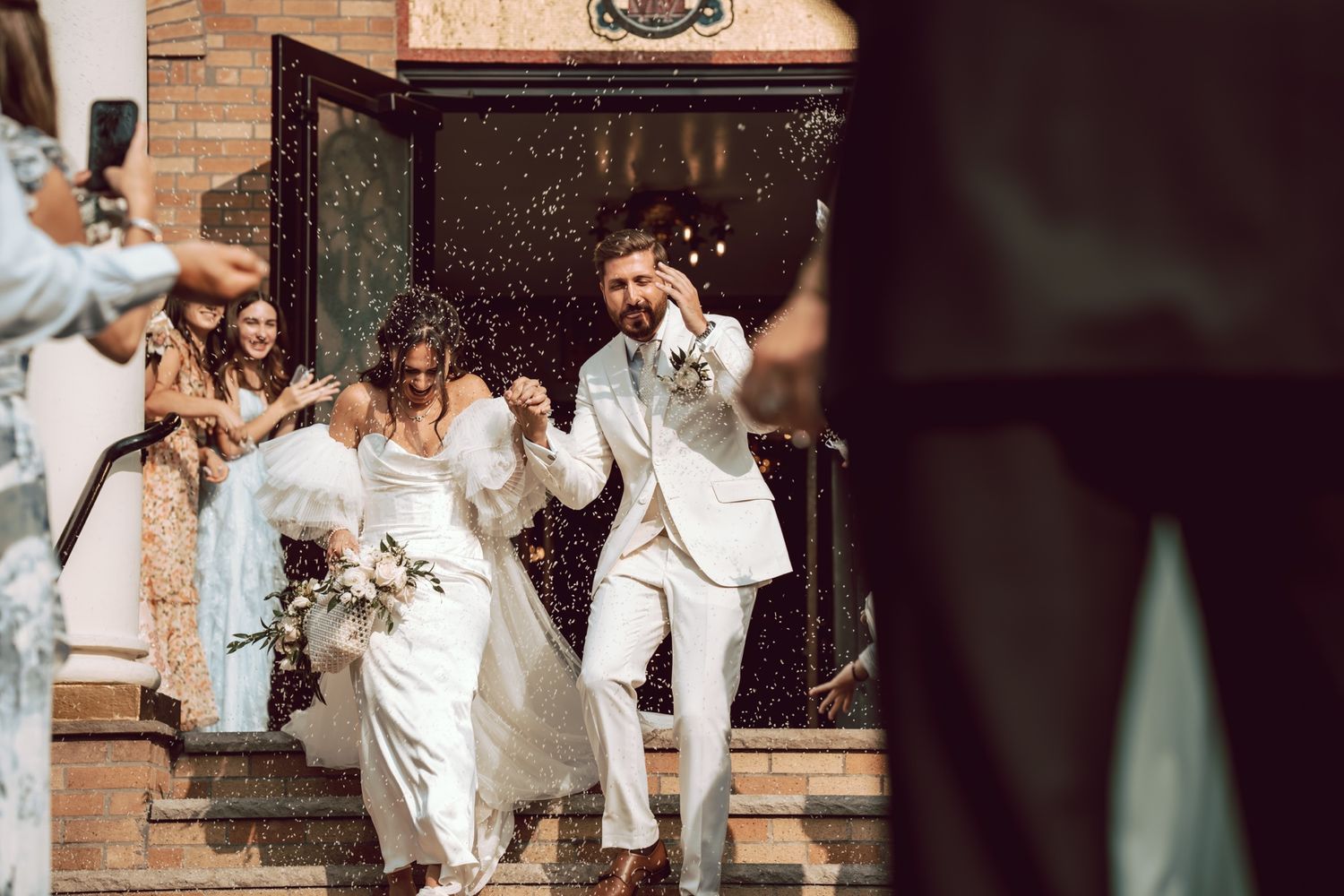 Joyful newlyweds exit church doors as guests celebrate with confetti shower, bride holding bouquet in elegant white gown.