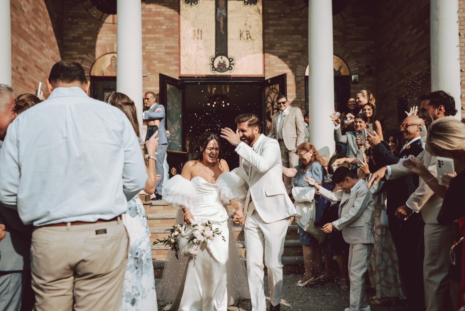 Smiling bride and groom walk through a cheering crowd of guests outside a church after their wedding.