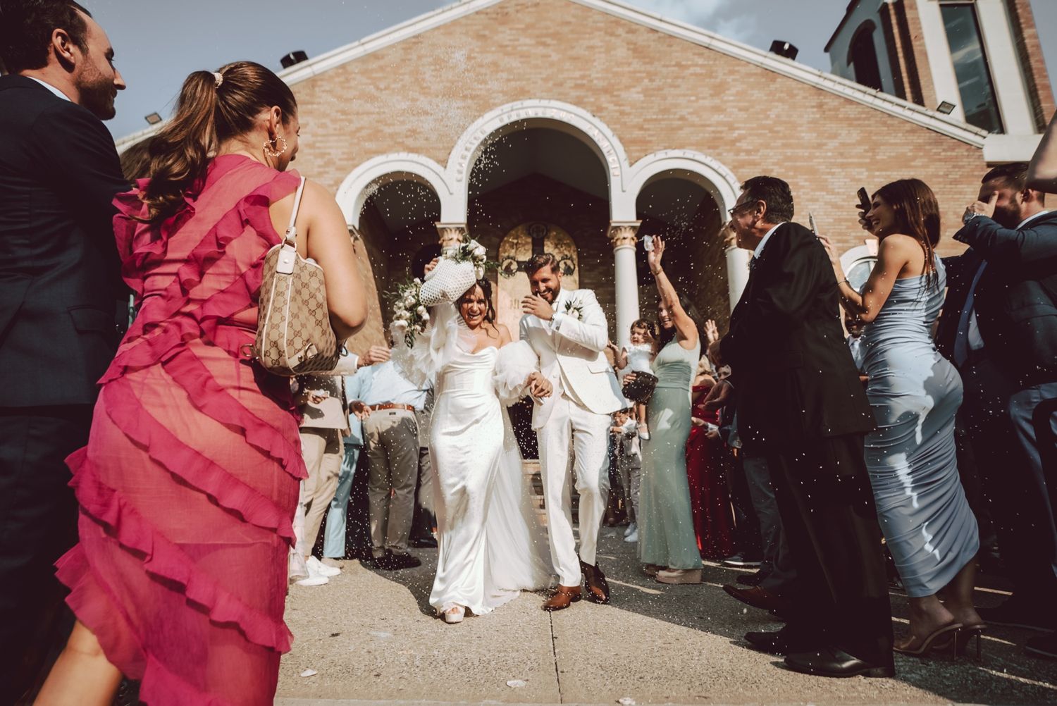 Newlyweds exit church amid cheering guests throwing confetti on sunny wedding day.