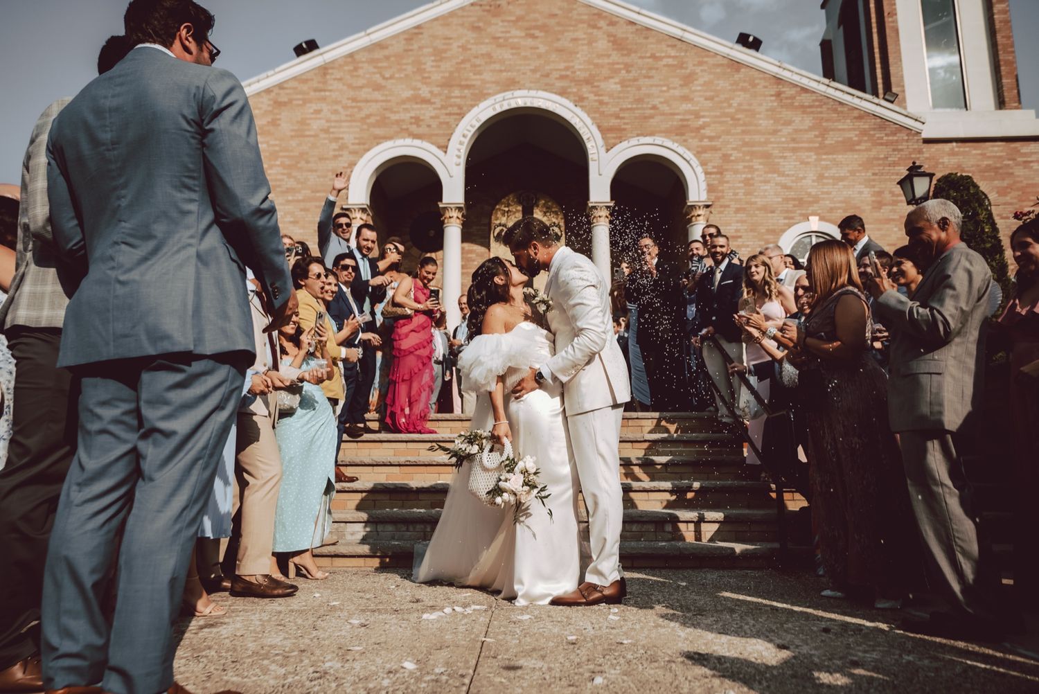 Bride and groom share kiss on church steps surrounded by celebrating wedding guests.