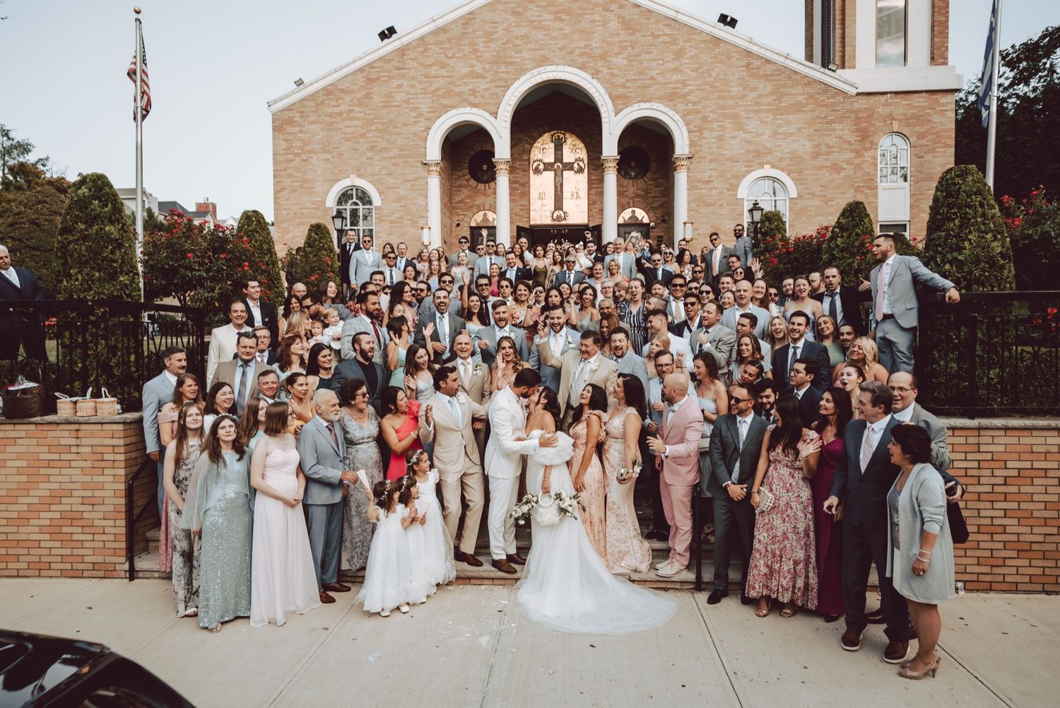 Wedding guests and bridal party pose on brick church steps for large group celebration photo.