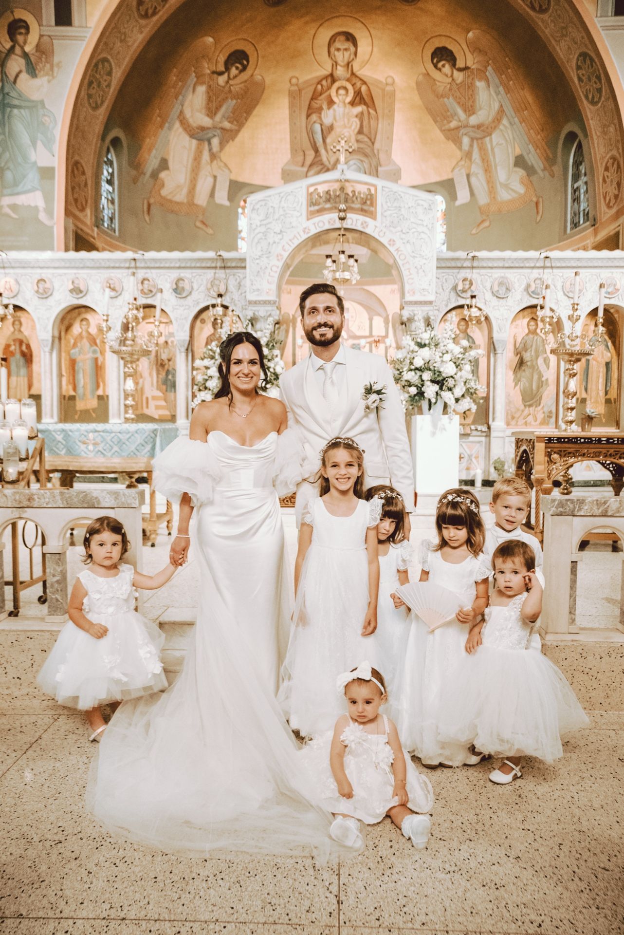 Bride and groom pose with flower girls in white dresses inside a decorated Orthodox church during their wedding ceremony.