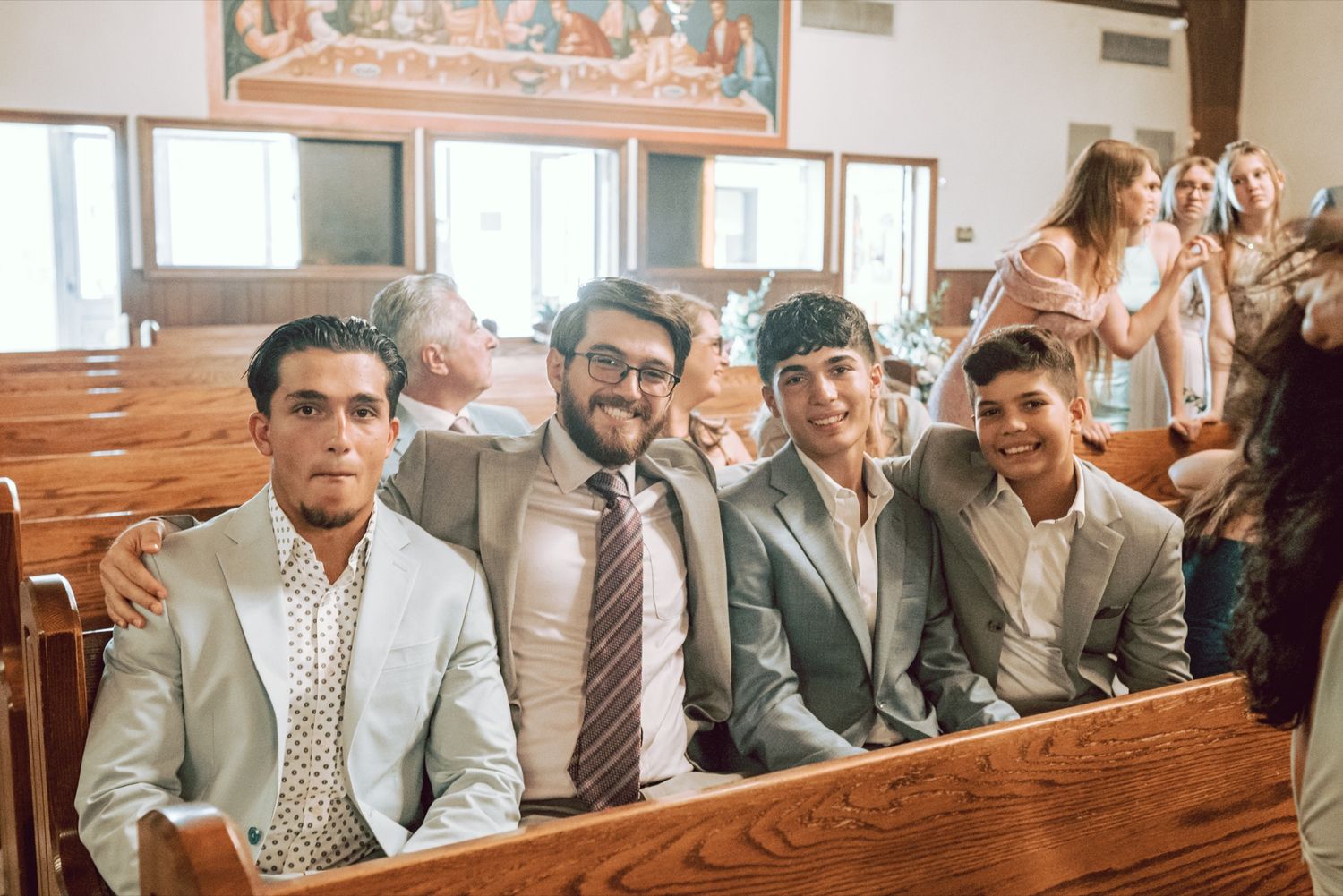 Four young men in suits smile while seated in wooden church pews during a wedding ceremony.