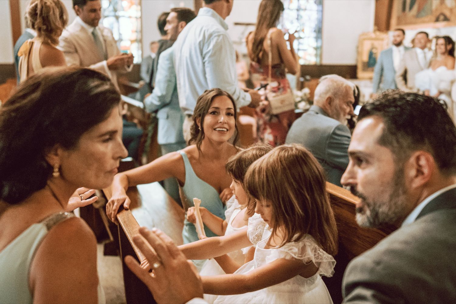 Guests socializing in church pews during a wedding ceremony, with a young girl in a white dress interacting with seated adults.